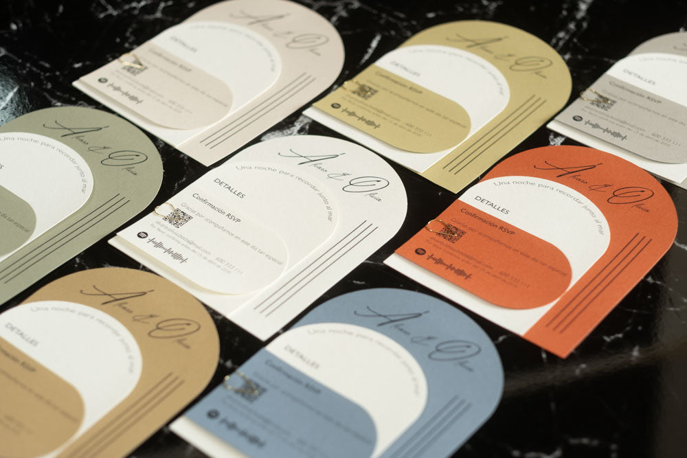 Colección de invitaciones de boda Aventurina en varios colores (terracota, verde, azul, miel y marfil), con diseño semicircular moderno y acabados en papel natural, dispuestas en mesa de mármol.