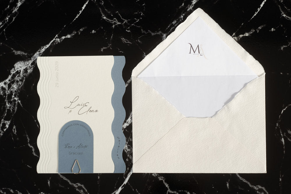 Invitación de boda Berilio en tonos azul y blanco, con diseño ondulado elegante y tarjeta adicional sujeta con clip dorado, acompañada de sobre artesanal blanco con forro interior personalizado.