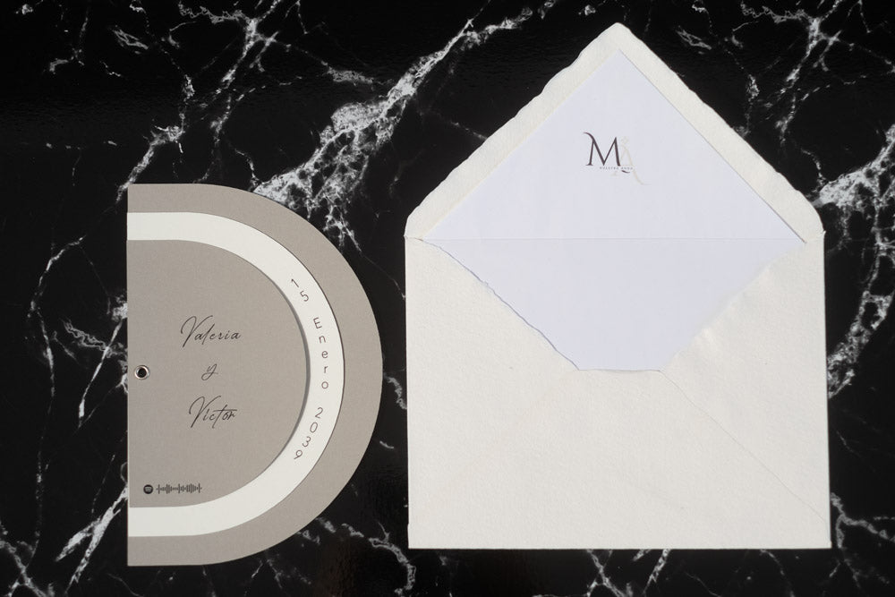 Invitación de boda modelo Cuarzo en color beige, semicircular cerrada, con los nombres de los novios y la fecha del enlace impresa en el borde exterior.
Con sobre blanco forrado.