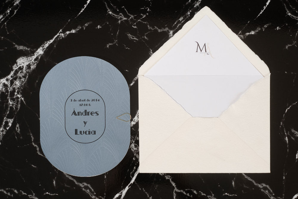 Invitación de boda ovalada en color azul con textura grabada y tipografía en negro, junto a sobre artesanal blanco con forro interior personalizado.