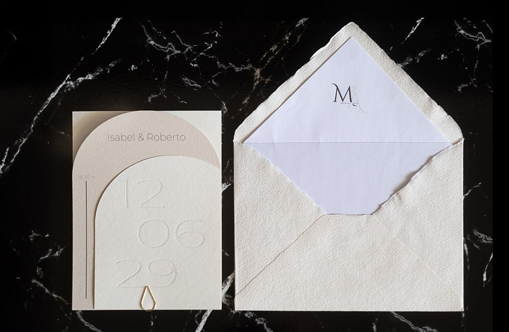 Invitación de boda semicircular en tono marfil con textura grabada y tipografía negra, acompañada de sobre artesanal blanco con forro interior personalizado.