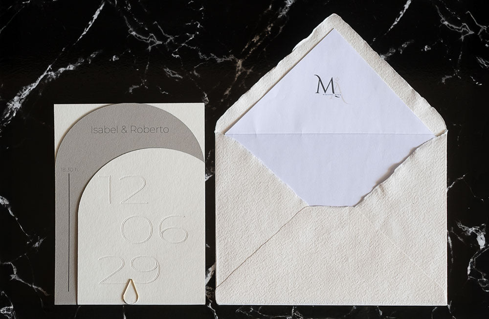 Invitación de boda semicircular en tono gris con textura grabada y tipografía negra, acompañada de sobre artesanal blanco con forro interior personalizado.