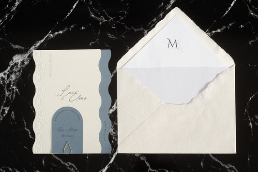 Invitación de boda Berilio en tonos azul y blanco, con diseño ondulado elegante y tarjeta adicional sujeta con clip dorado, acompañada de sobre artesanal blanco con forro interior personalizado.