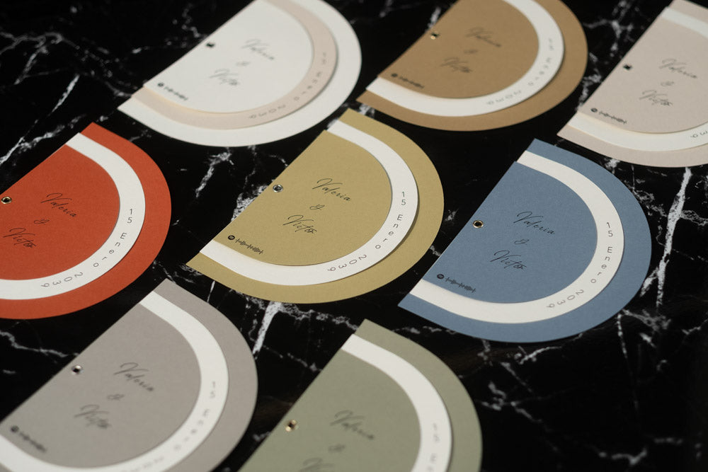Vista superior de varias invitaciones de boda modelo Cuarzo en diferentes colores (terracota, oliva, azul, arena, beige y gris), con diseño semicircular moderno.