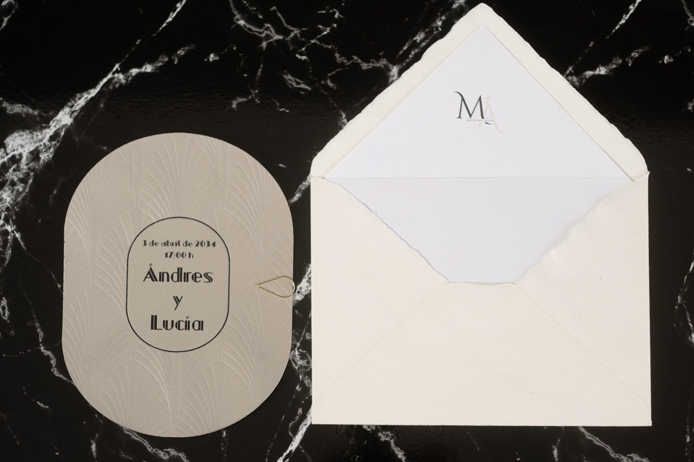 Invitación de boda ovalada en tono gris claro con textura grabada y tipografía negra, acompañada de sobre artesanal blanco con forro interior personalizado.