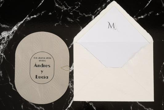 Invitación de boda ovalada en tono gris claro con textura grabada y tipografía negra, acompañada de sobre artesanal blanco con forro interior personalizado.