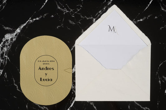 Invitación de boda ovalada en color dorado con textura grabada y tipografía negra, acompañada de sobre artesanal blanco con forro interior personalizado.