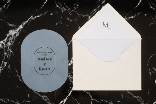 Invitación de boda ovalada en color azul con textura grabada y tipografía en negro, junto a sobre artesanal blanco con forro interior personalizado.