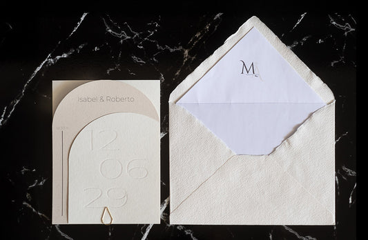 Invitación de boda semicircular en tono marfil con textura grabada y tipografía negra, acompañada de sobre artesanal blanco con forro interior personalizado.