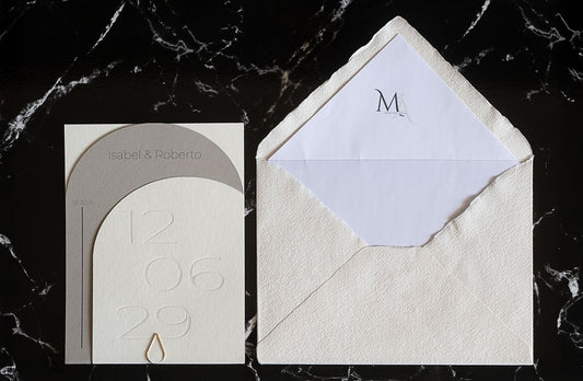 Invitación de boda semicircular en tono gris con textura grabada y tipografía negra, acompañada de sobre artesanal blanco con forro interior personalizado.