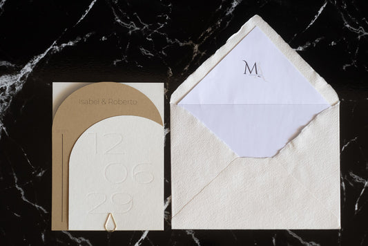 Invitación de boda semicircular en tono arena con textura grabada y tipografía negra, acompañada de sobre artesanal blanco con forro interior personalizado.