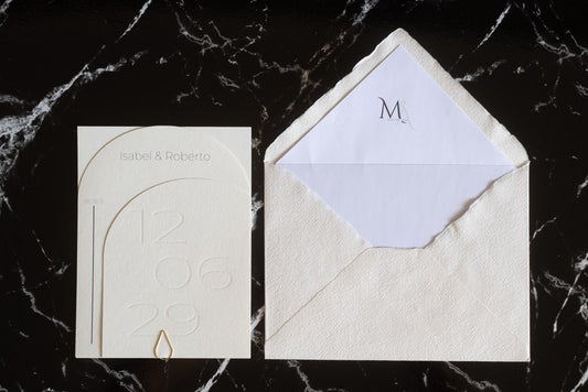 Invitación de boda semicircular en tono nieve con textura grabada y tipografía negra, acompañada de sobre artesanal blanco con forro interior personalizado.